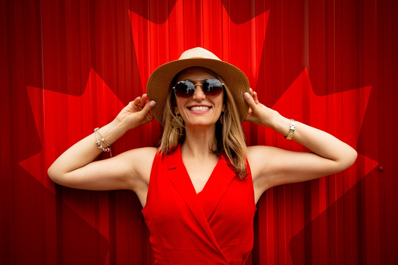 A fashionable woman in a red dress and hat smiling proudly in front of a Canadian flag backdrop.